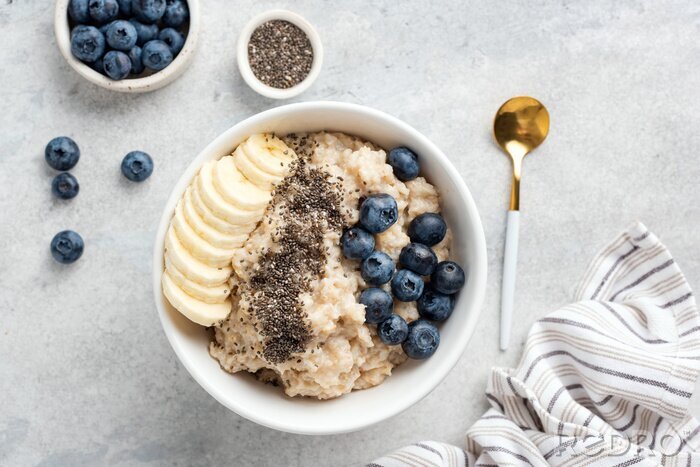 Poster Breakfast oatmeal porridge with banana, blueberries, chia seeds on grey concrete background. Table top view of healthy food for weight loss, clean eating, vegan and vegetarian diet