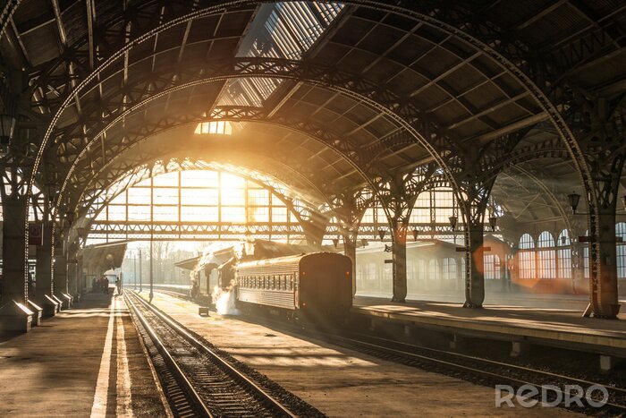 Poster Old railway station with a train and a locomotive on the platform awaiting departure. Evening sunshine rays in smoke arches.